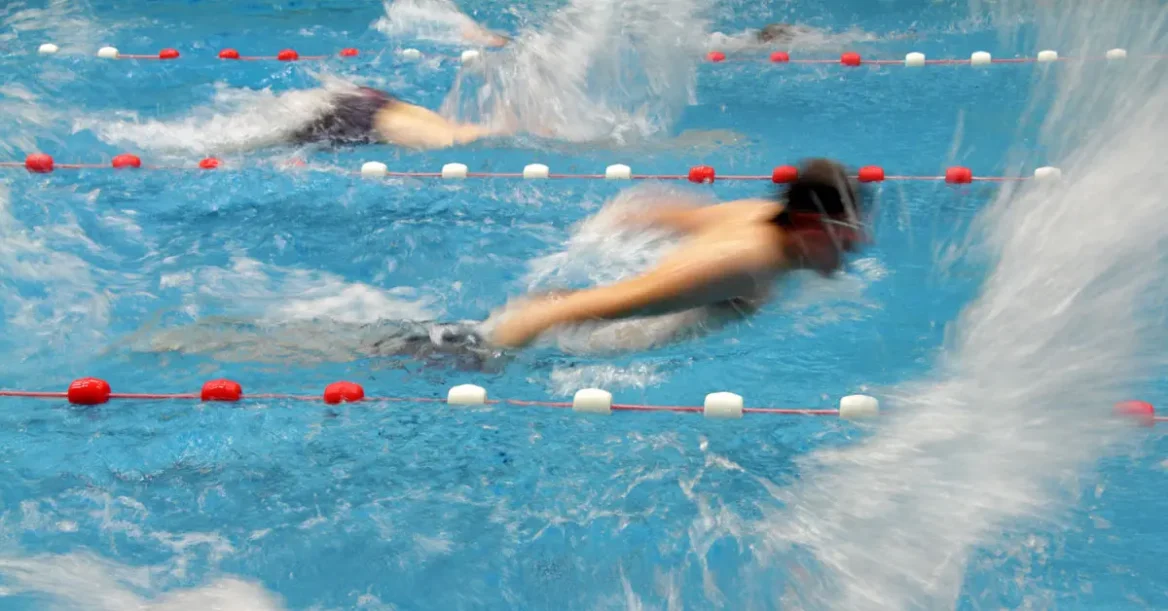An action shot of a man swimming laps in a commercial swimming pool. Other swimmers in adjacent lanes create water splashes in the fore- and background.